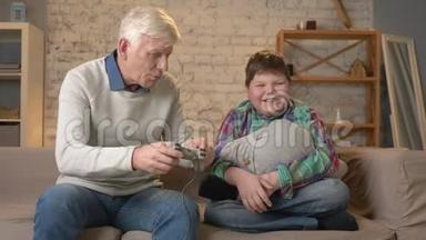 爷爷和孙子坐在沙发上玩游戏机。 一位老人坐在沙发上